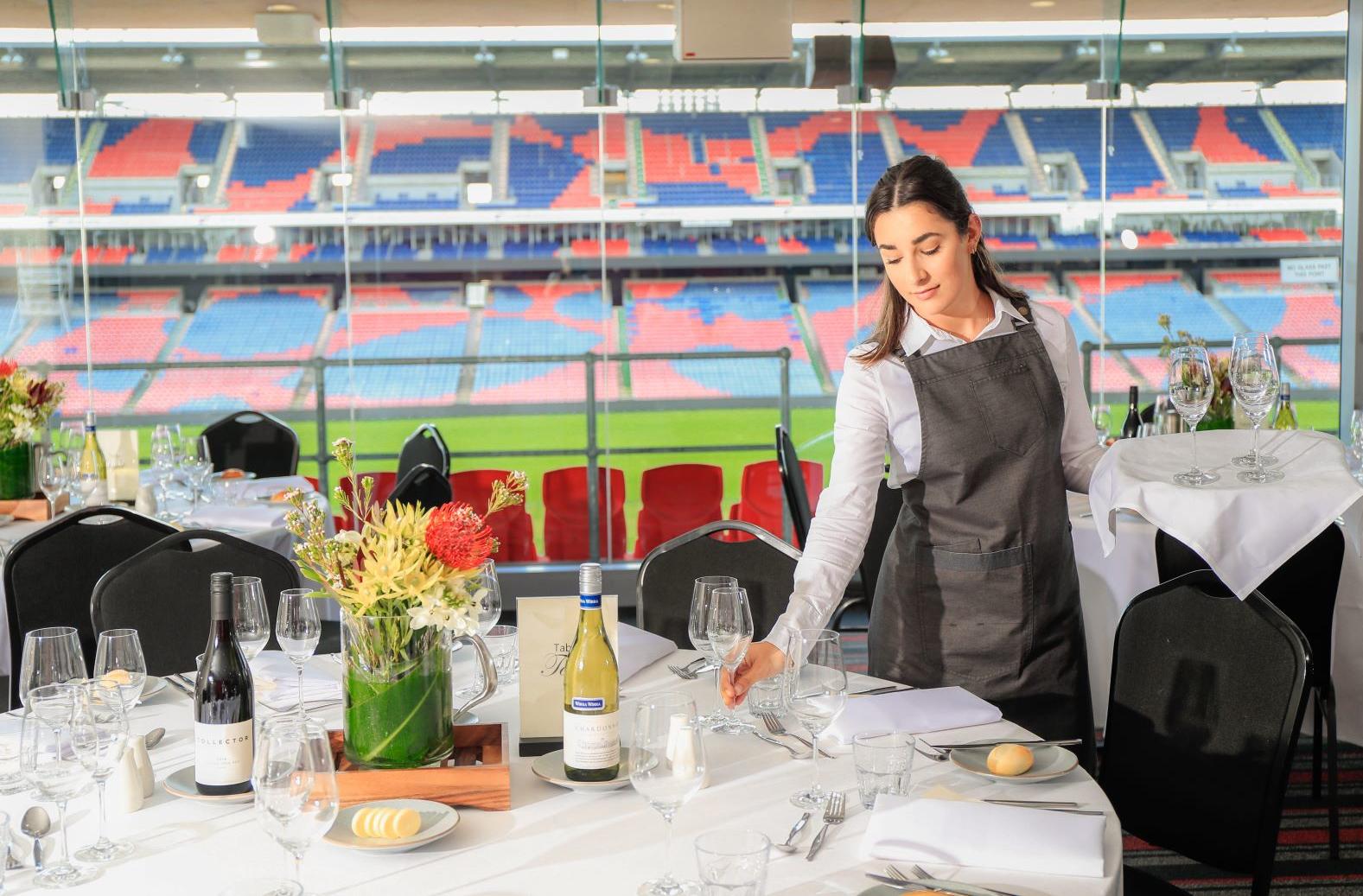 Image of a banquet table, dressed for a formal meal including a native floral centerpiece. The background shows the view of the Stadium through the window. A Service Staff member is holding a bar tray with wine glass and is placing one on the table