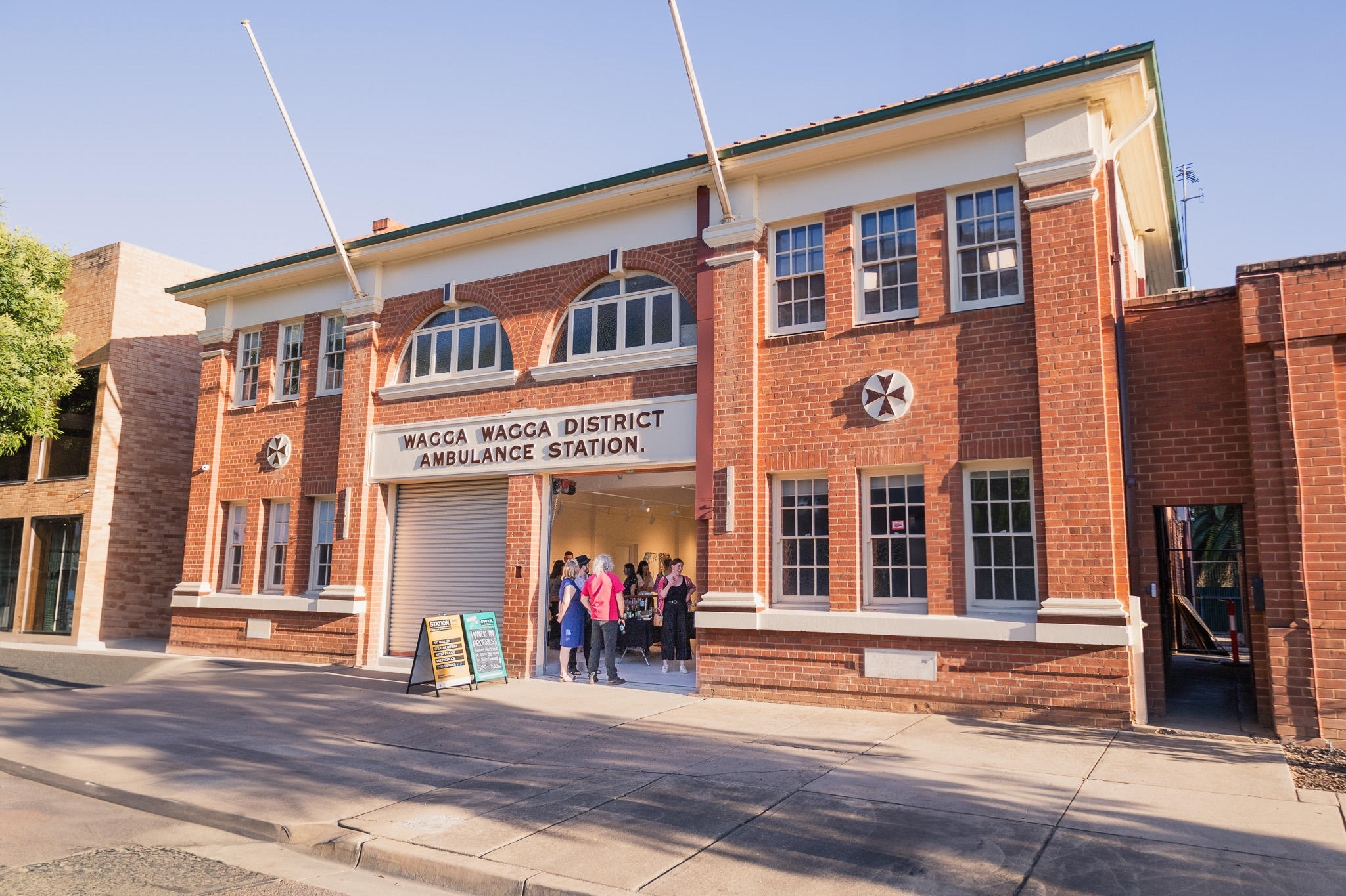 Photo of the front of Station Creative Workspace. The building is a historic ambulance station with two roller doors in the centre of the front, one of them is raised to reveal people inside mingling.